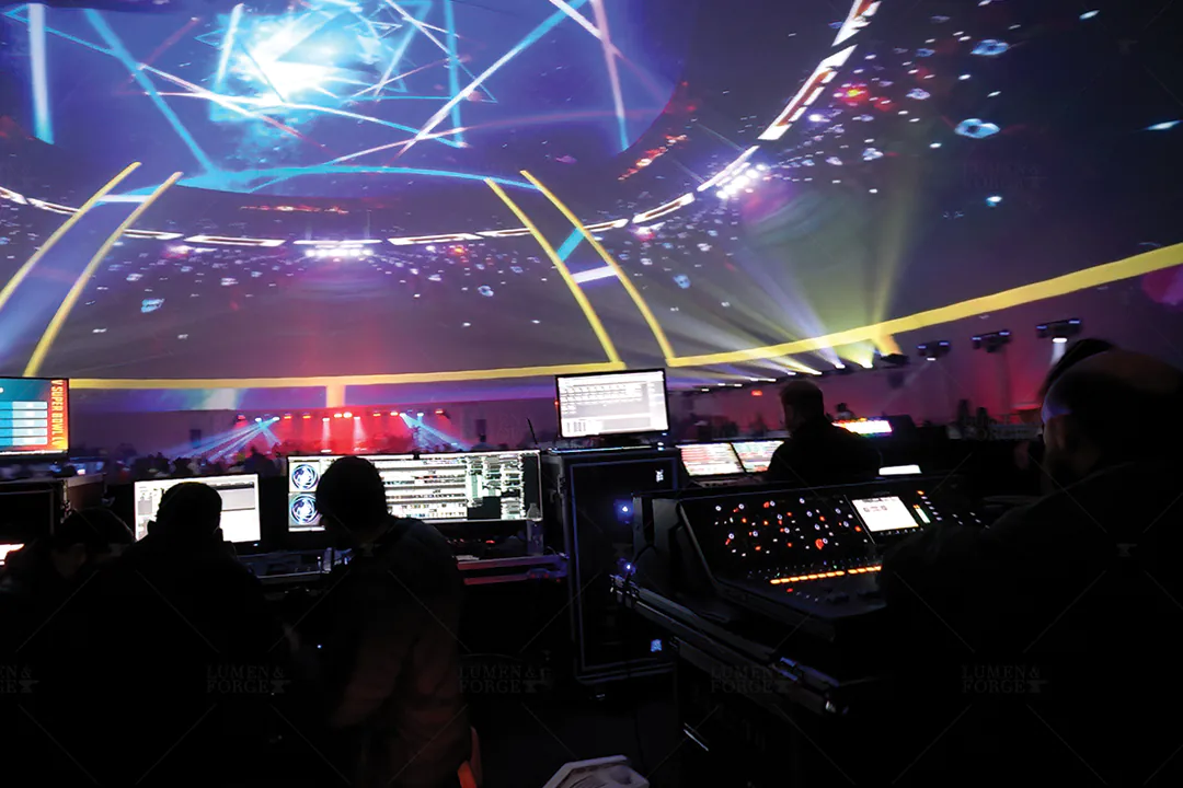 Show crew at lighting and media consoles beneath the Atlanta Super Bowl dome, lit with blue projected light patterns