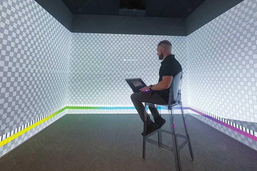 Man on a stool with a laptop calibrating a 360 projection room, checkerboard test pattern mapped on every wall