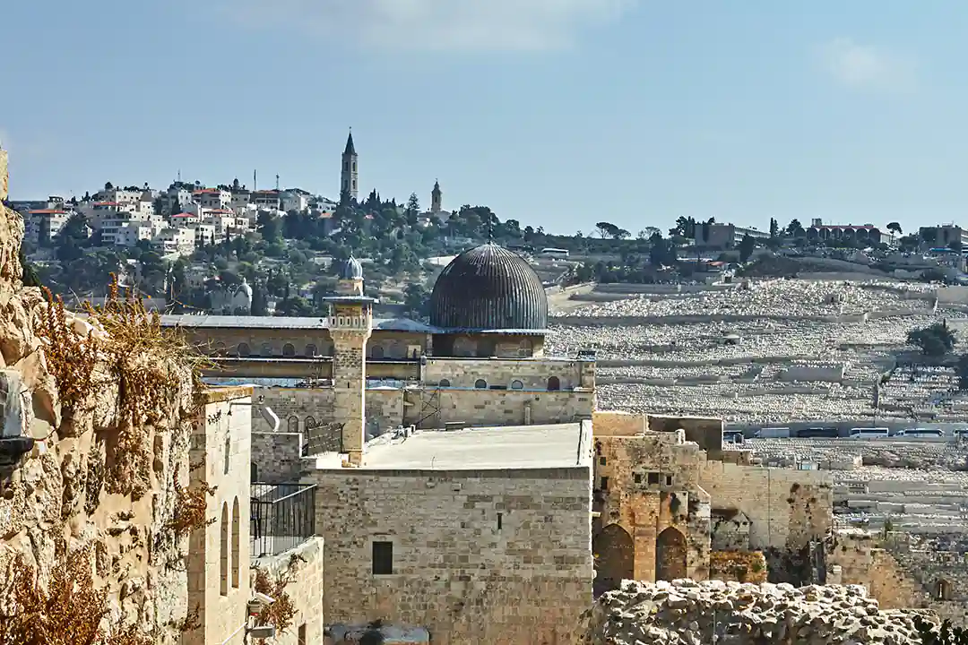 Al-Aqsa Mosque with its dark grey dome and stone minaret, hillside neighborhood and cemetery behind