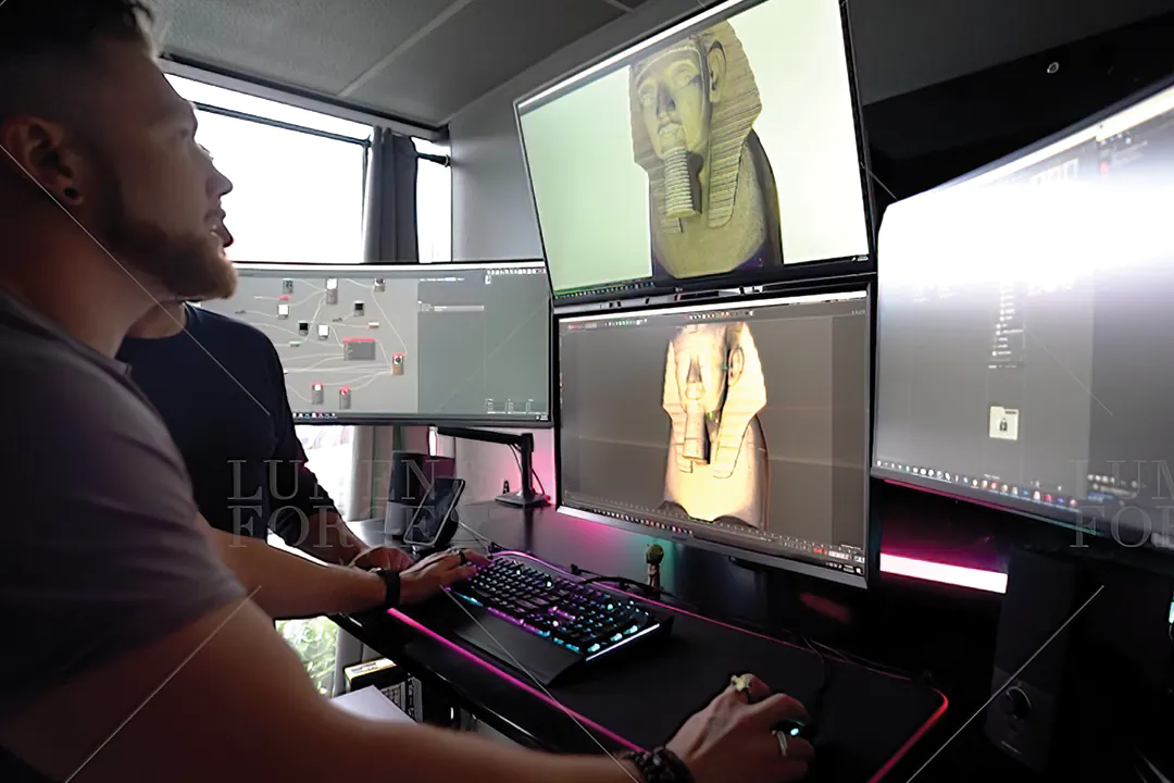 Two 3D artists sculpting an Egyptian pharaoh head model across several monitors at a studio workstation