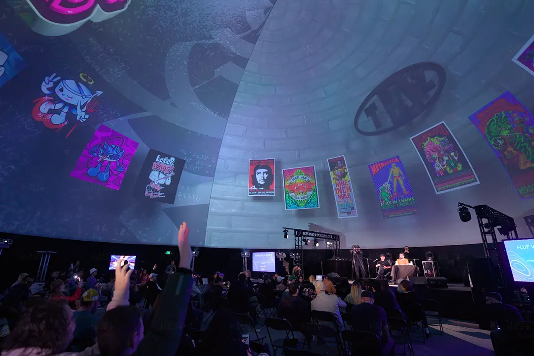 Audience inside a 360 dome watching a stage panel beneath projected poster art, one attendee raising a hand