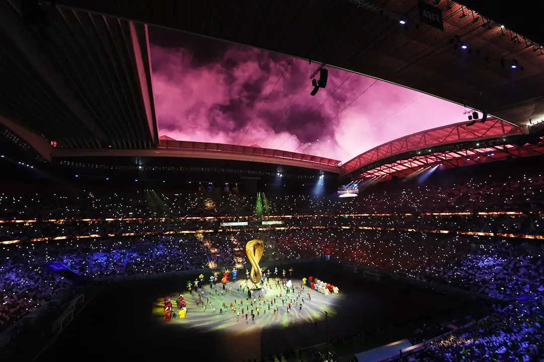 FIFA World Cup opening ceremony in Qatar, performers around a giant golden trophy on a lit pitch with pink smoke above the roof