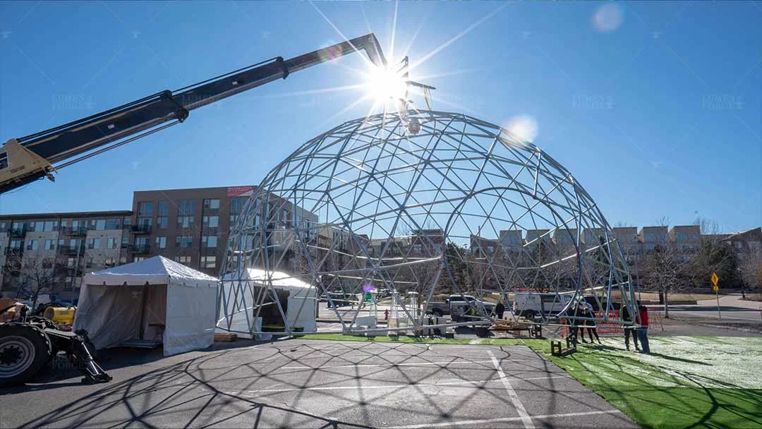 Crane assembling a 44 ft geodesic dome metal frame for Capital One in a sunny parking lot beside white tents