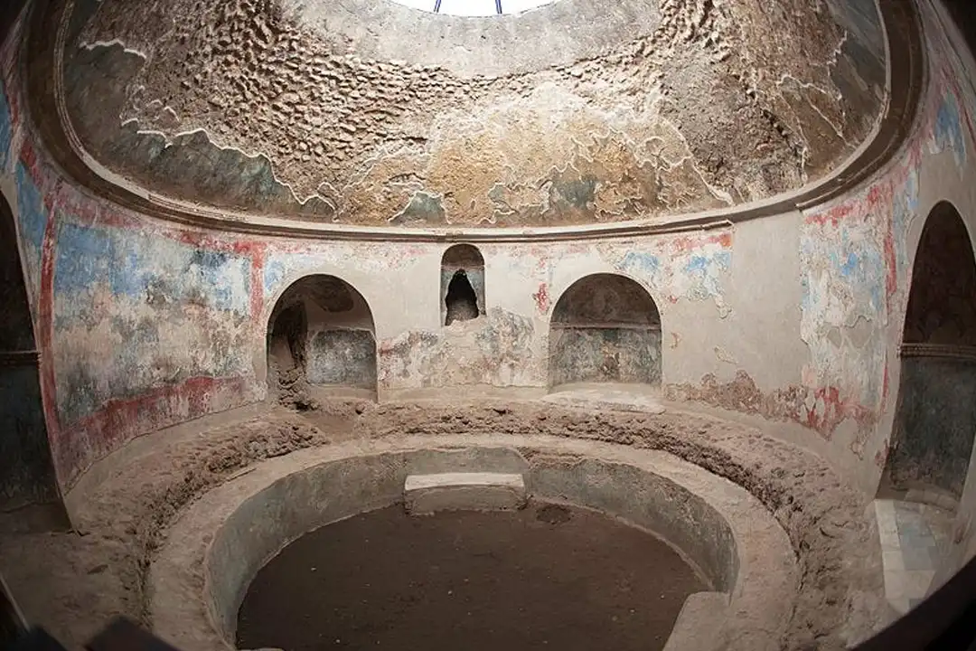 Ancient domed bathhouse room with faded frescoes, arched niches and a round plunge pool lit by an opening in the dome