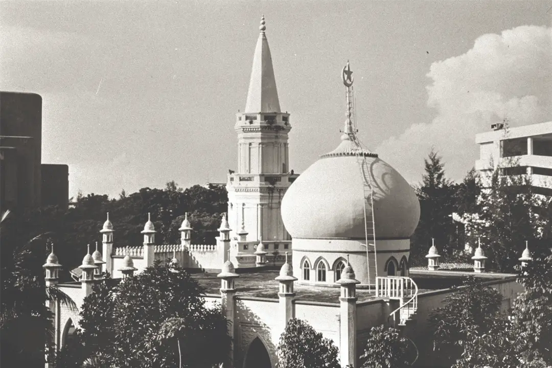 Black and white photo of a mosque with a white onion dome, crescent finial and tall minaret among trees
