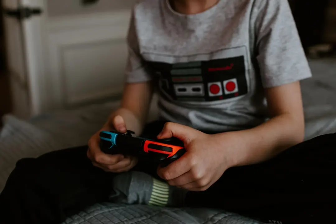 Child sitting on a bed playing a Nintendo Switch with a Joy-Con grip, wearing a retro gamepad T-shirt