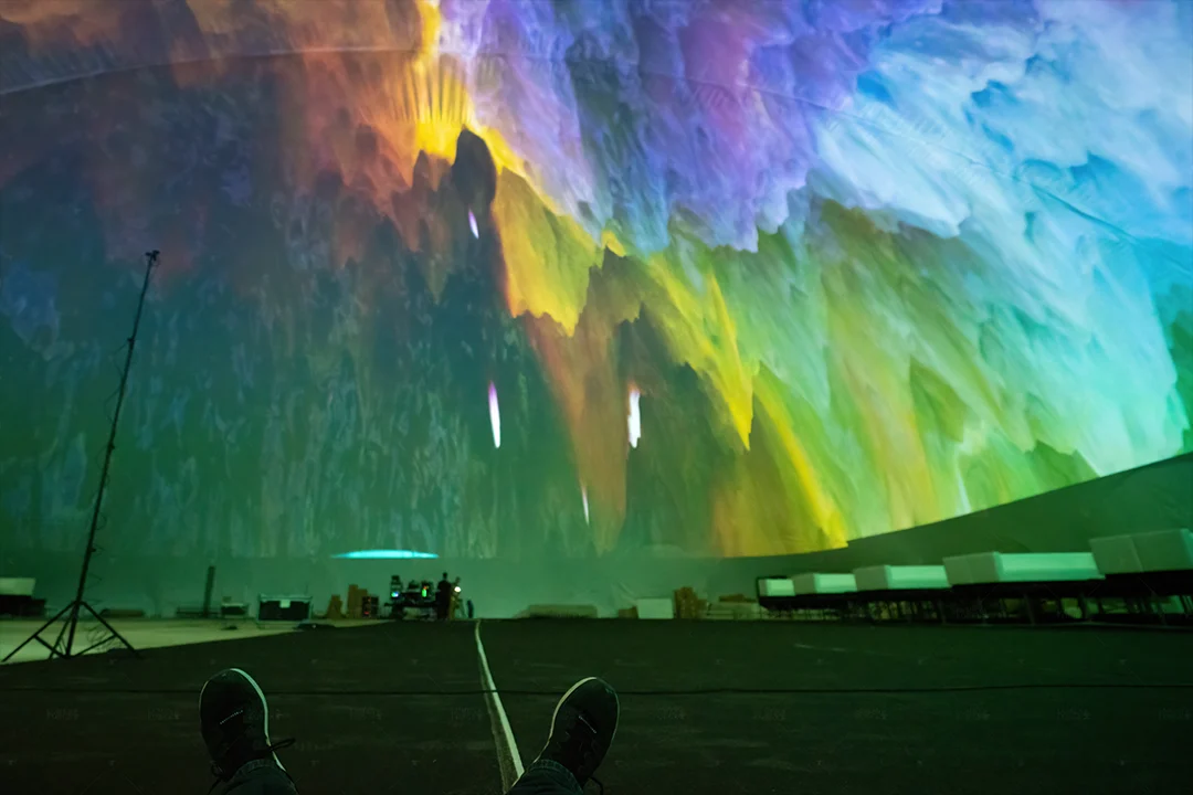 Point-of-view shot of a reclining viewer's feet under a colorful smoke-like projection in the Miami dome
