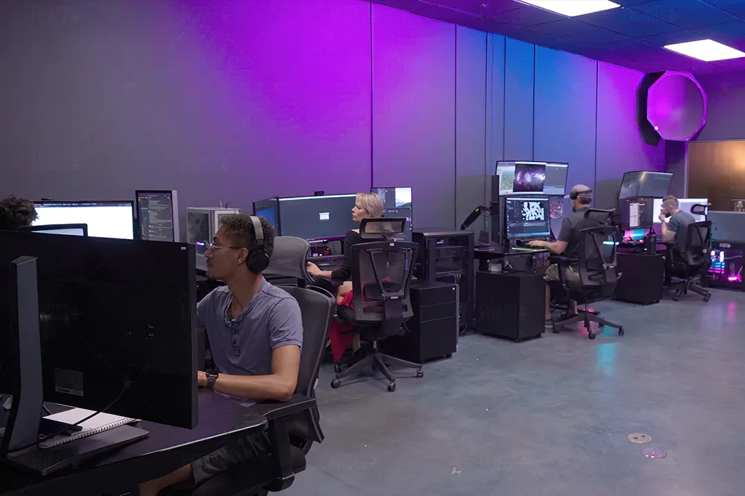 Artists at multi-monitor workstations in the Lumen and Forge video game content production office under purple light