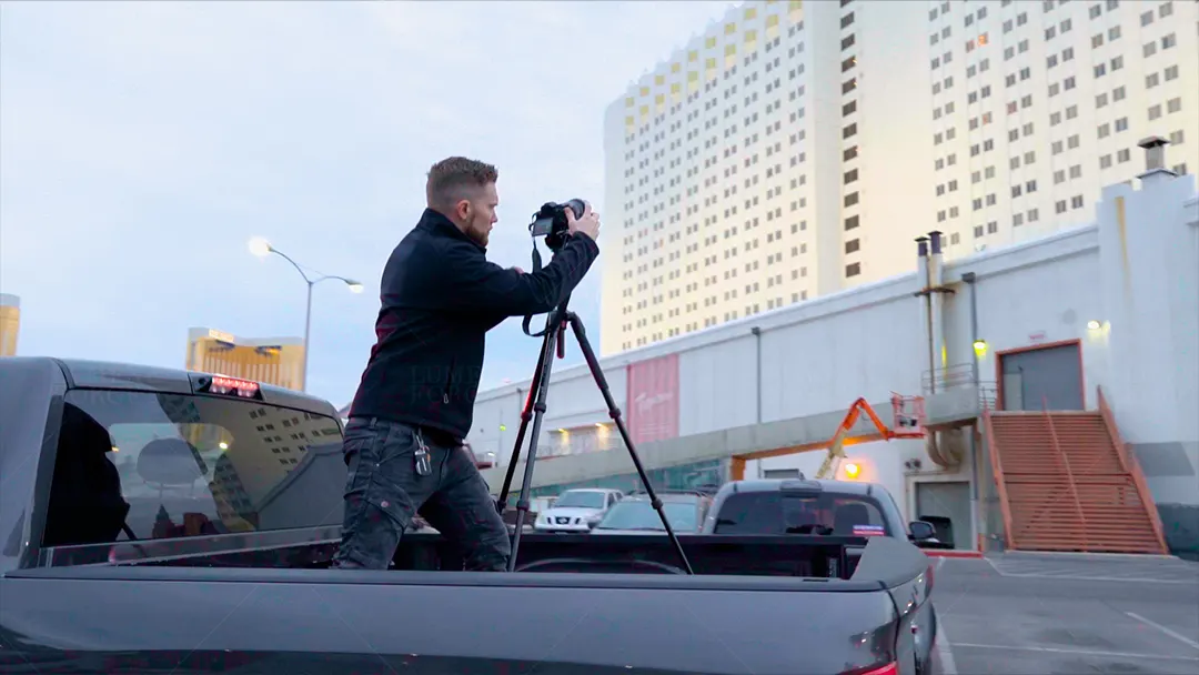 Photographer standing in a pickup truck bed aiming a tripod-mounted camera at the Tropicana hotel tower