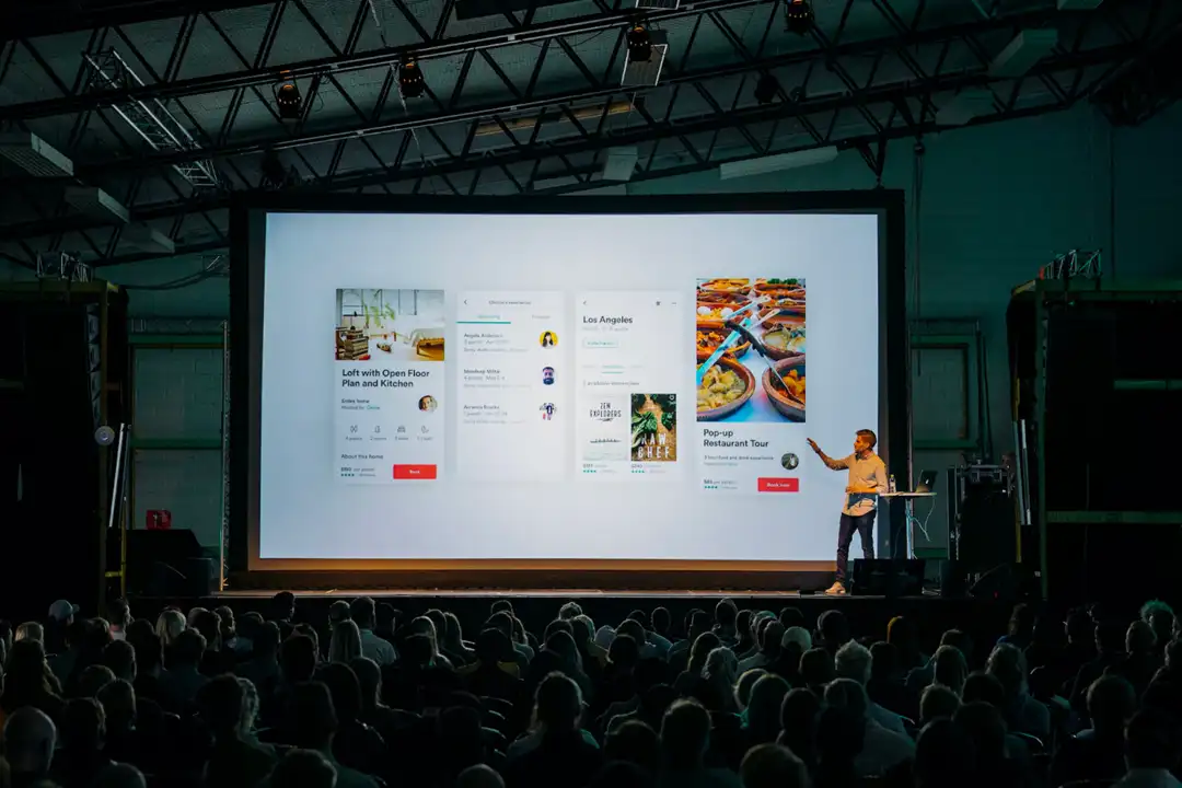 Speaker presenting app screens on a large projection screen to a packed audience in a warehouse venue