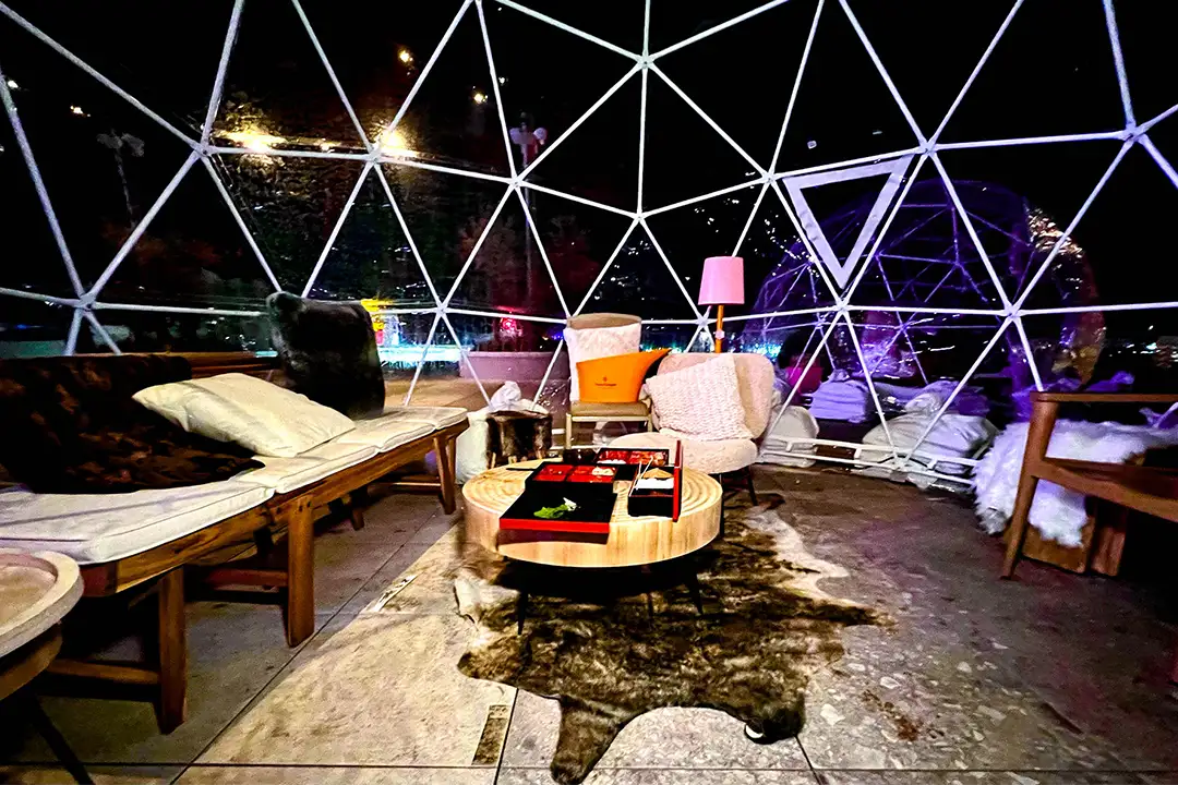 Inside a geodesic dome at night with cushioned benches, a cowhide rug and a round table, and a purple-lit igloo beyond