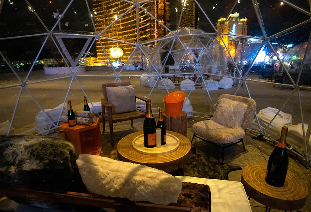 Inside a Rose Rooftop igloo at night with champagne bottles on wood tables, fur cushions and lit Strip towers outside