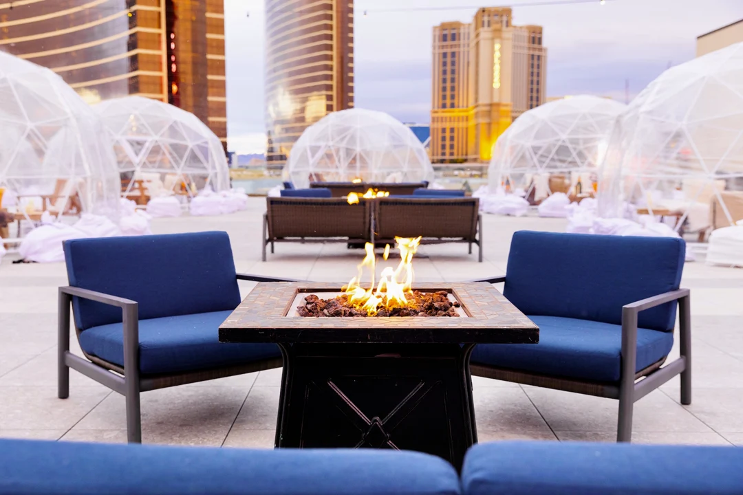 Fire pit ringed by blue lounge chairs on the Resorts World rooftop, with clear igloo domes and Las Vegas towers behind