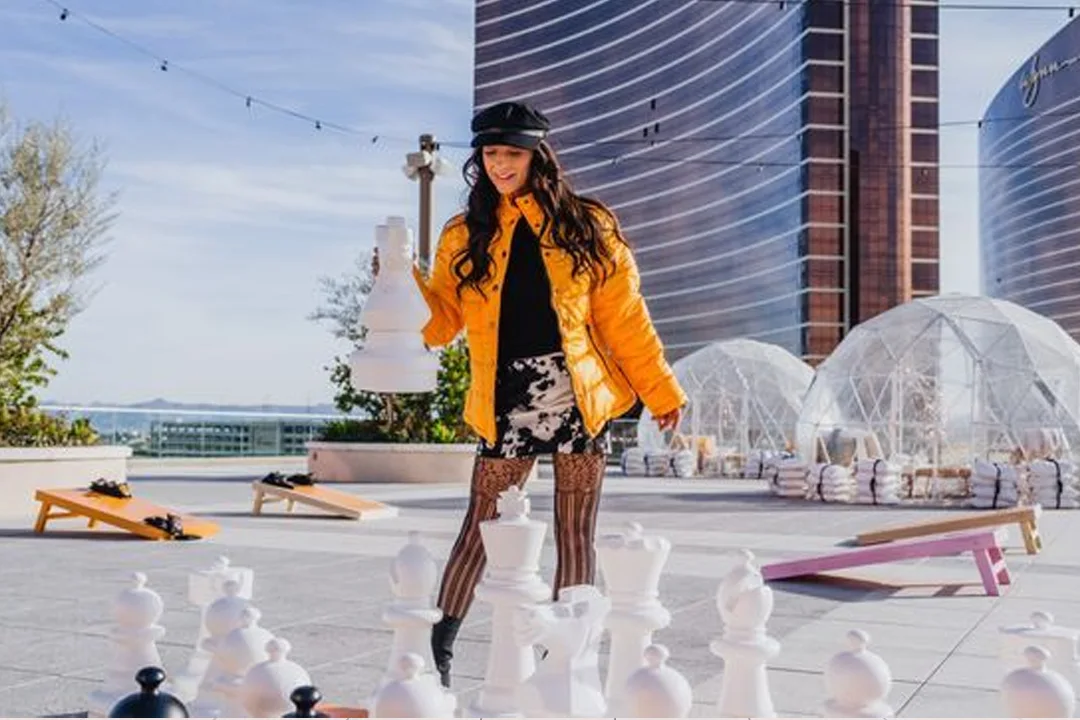 Woman in a yellow puffer jacket lifting a piece on a giant rooftop chess set, with igloo domes and cornhole boards behind