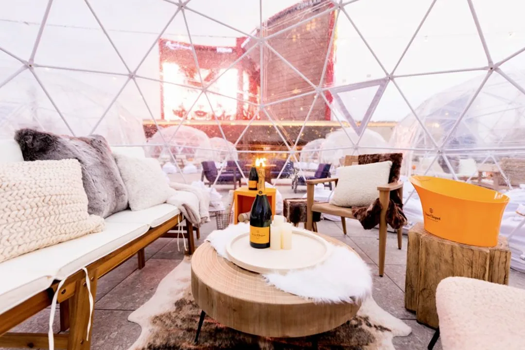 Inside a clear igloo dome at Resorts World with faux fur pillows, a wood table, champagne and a yellow ice bucket