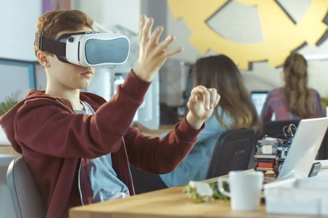 Student wearing a VR headset reaching out with both hands in a classroom, classmates working at computers behind him
