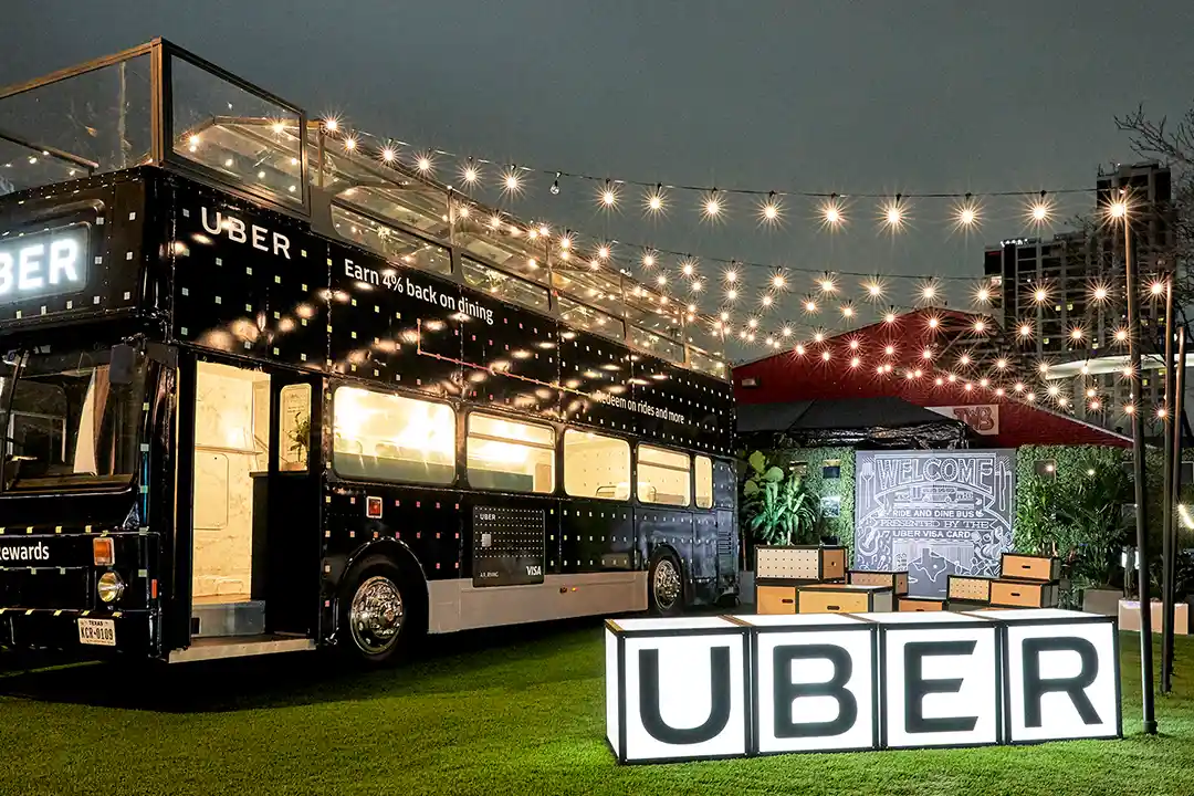 Uber-branded double-decker bus under string lights with illuminated UBER letter cubes at a New York City activation