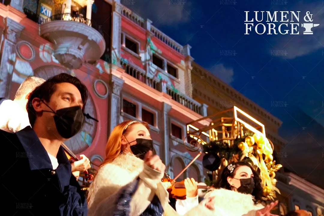 Holiday performers in face masks, one with a violin, in front of the projection-mapped clock tower at The Venetian