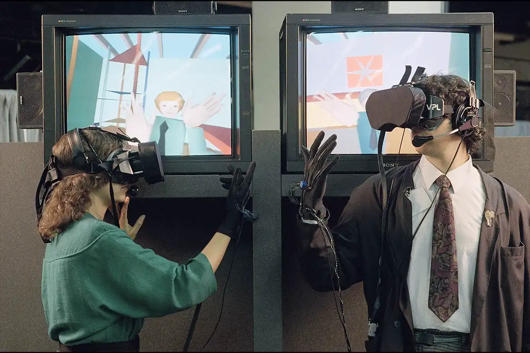 Two people in VPL headsets and data gloves gesturing before monitors showing a simple 3D virtual world with an avatar