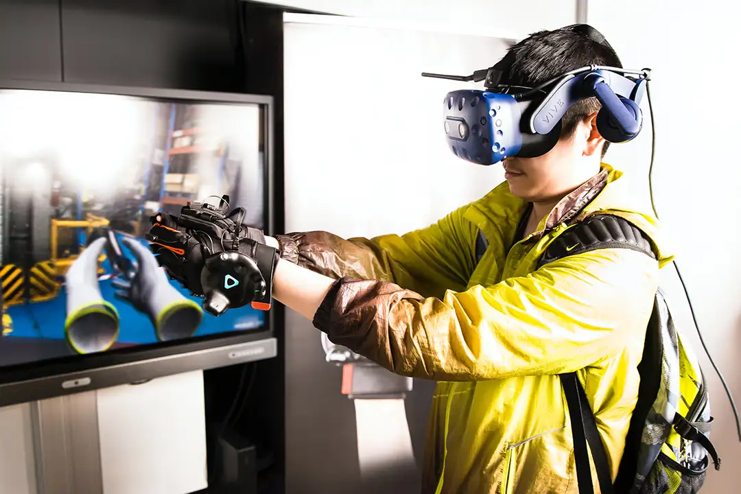 Man in a Vive headset and haptic gloves reaching out as a screen mirrors his virtual hands, shown at XR Expo 2019