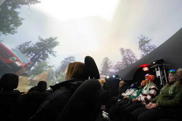Audience in winter hats seated in the Jeep 4D theater dome, watching a red Jeep drive a forest trail on the curved screen