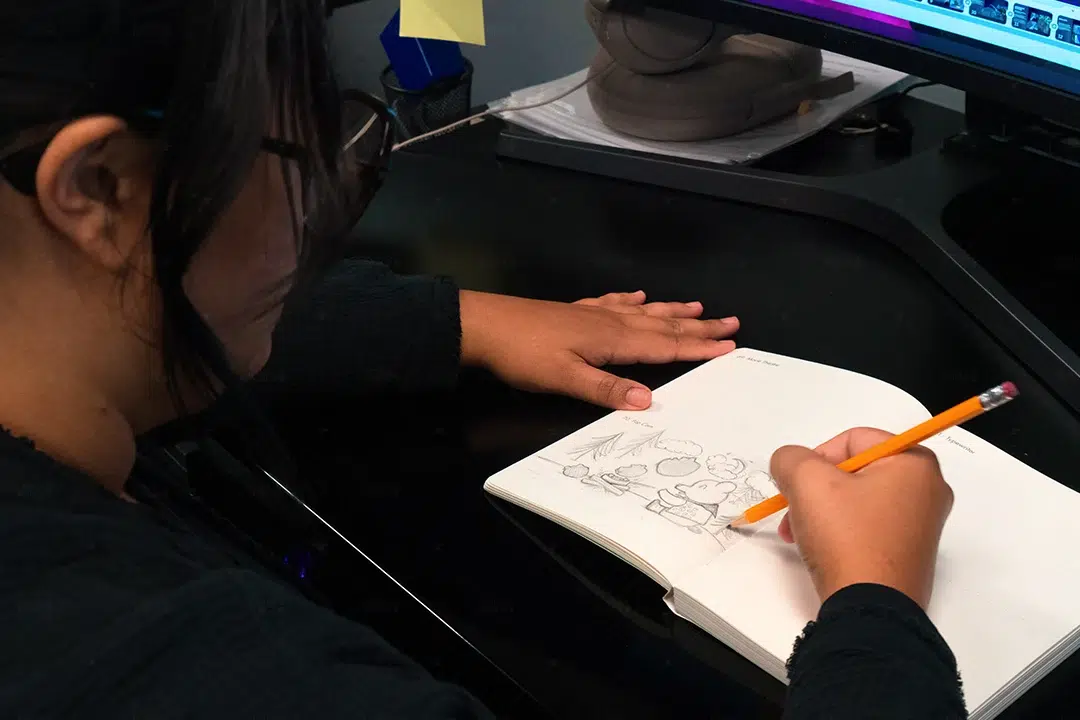 Animator sketching cartoon characters in a notebook with an orange pencil at a desk