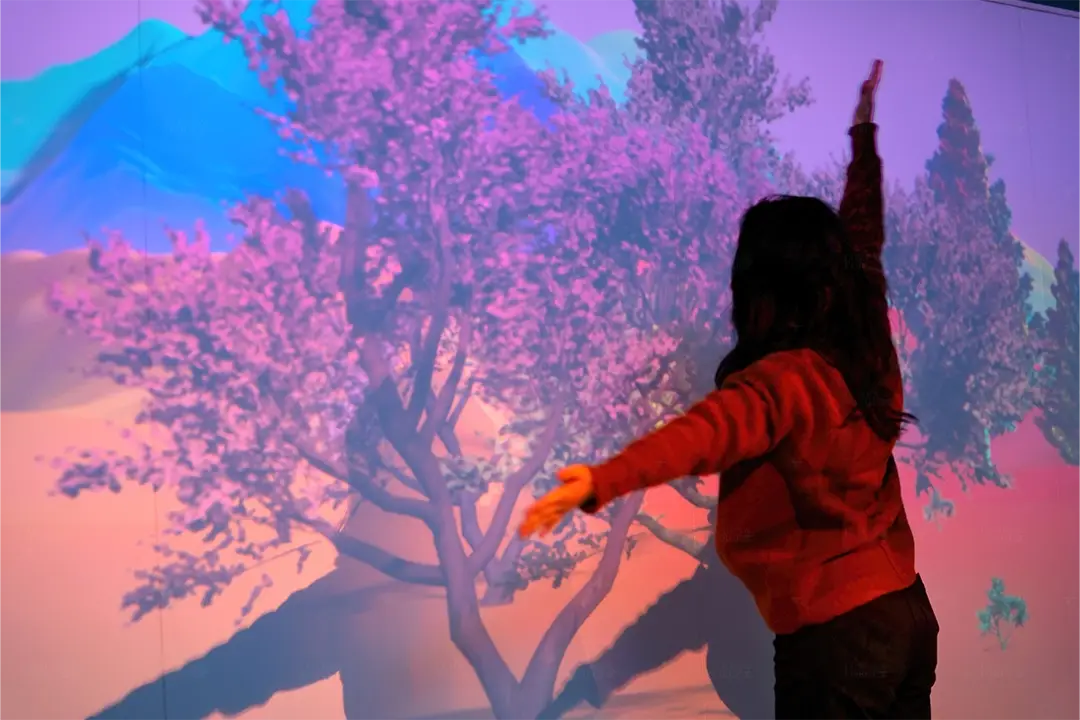 Child in a red sweater raising her arms before a projected pink blossom tree in an interactive immersive room