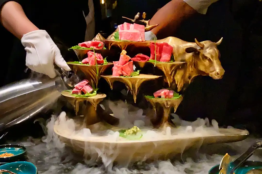 Server pouring dry ice fog over a gold bull sculpture stand holding tiers of raw sliced beef at The X Pot Las Vegas
