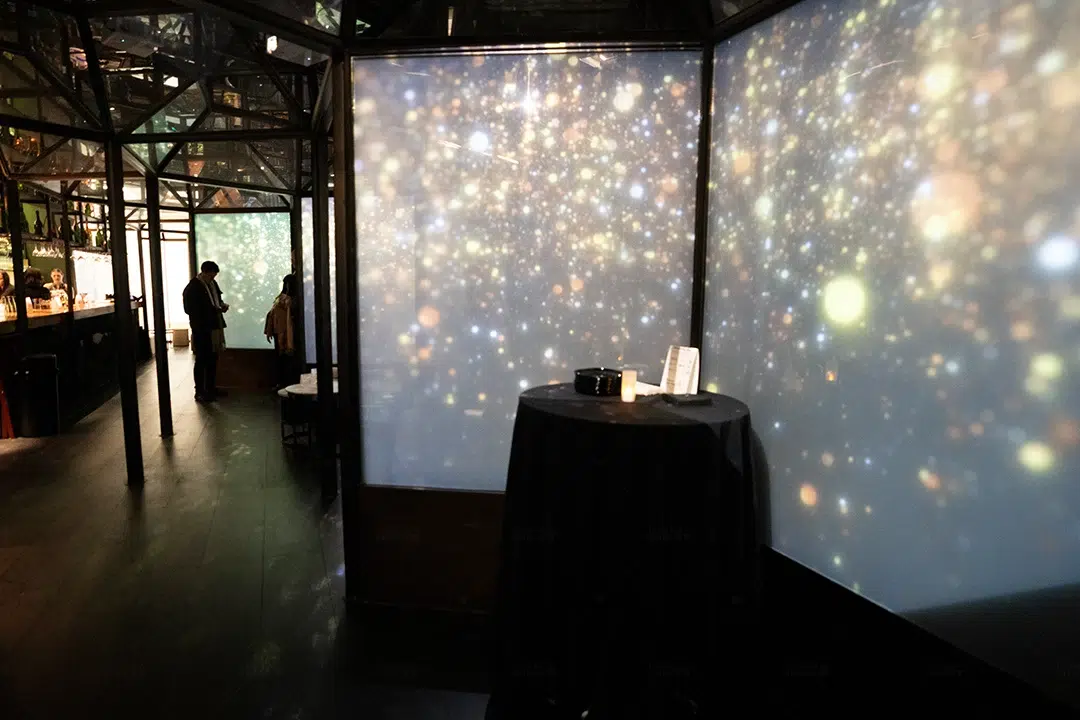 Glass-walled room at The X Pot glowing with projected starry light, a black-draped cocktail table in front