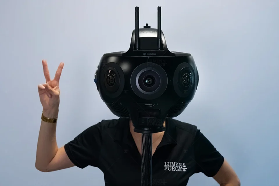 A person hides their head behind a Titan 360 camera to give the illusion the person has a giant camera-head. They are posing with a two-finger peace sign looking like a cyborg-droid.