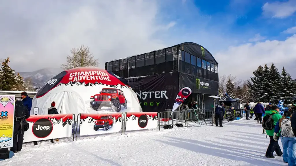 A Jeep-branded geo dome showcasing a 360-degree immersive experience at the Winter X Games, with Monster Energy's activation booth nearby, set against a snowy mountain backdrop.