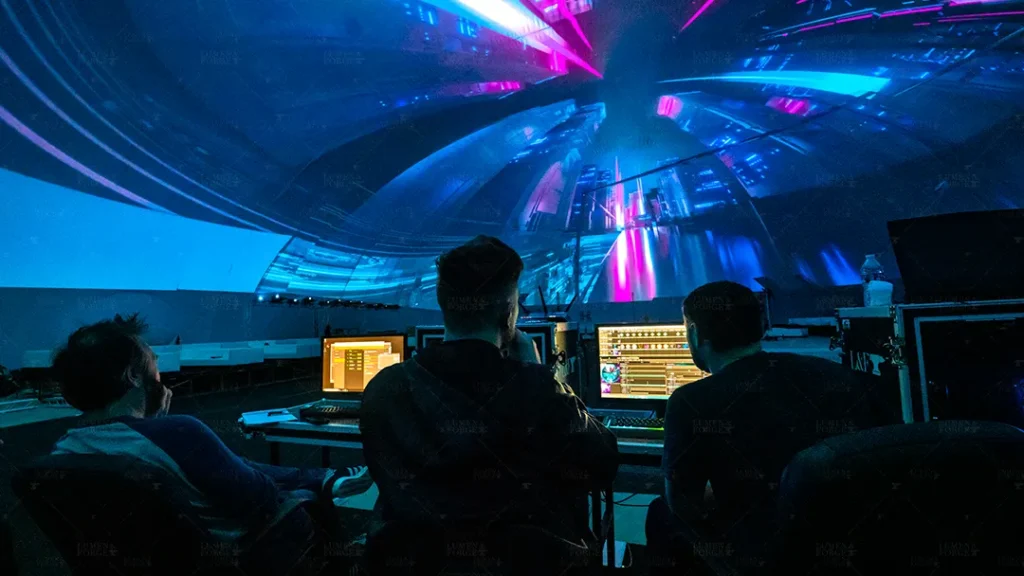 A group of technicians working inside Miami's largest 360-degree projection dome, focused on programming and testing the vibrant, immersive visuals displayed across the dome's interior surface. The image captures the intense blue and pink hues of the digital graphics, creating a futuristic atmosphere.