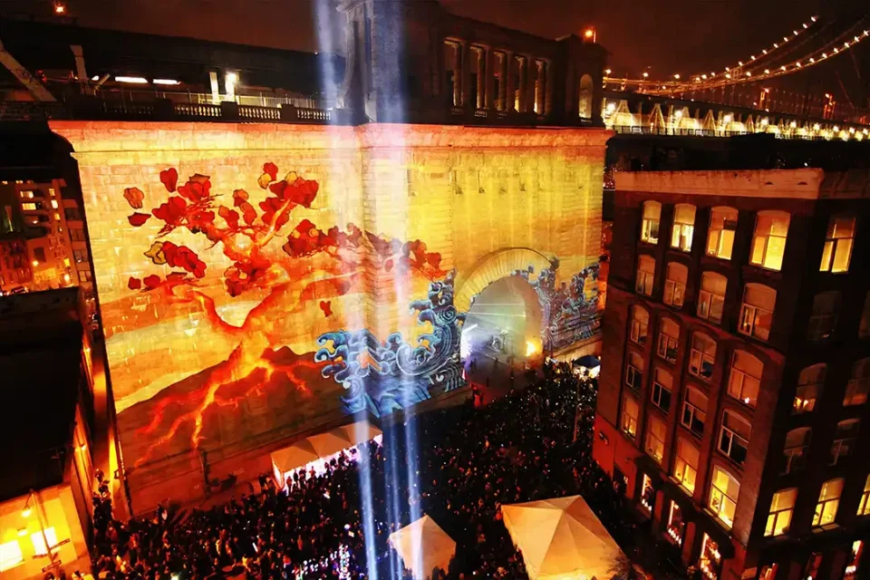 A vibrant projection mapping display illuminates a historic brick building in New York City, featuring dynamic red and blue imagery of a tree and waves. The visual spectacle draws a large crowd of onlookers, showcasing the transformative power of projection mapping in an urban setting.