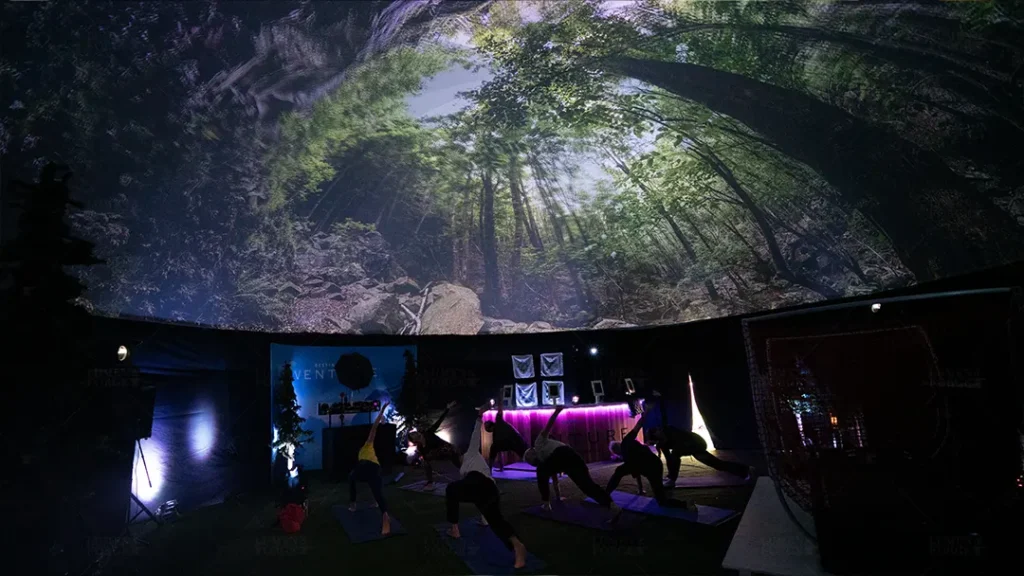 An immersive yoga session takes place under a giant projection dome in Denver, Colorado. The dome ceiling is covered with a lush, high-definition projection of a forest canopy, creating a serene natural atmosphere. People in the foreground are engaged in yoga poses, illuminated by the calming glow of the projection.