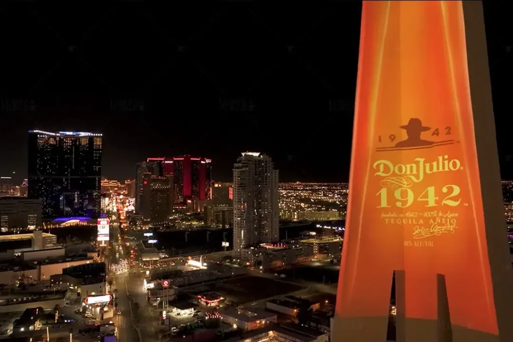 A large-scale projection mapping of the Don Julio 1942 tequila logo displayed on the Stratosphere Tower in Las Vegas at night. The illuminated branding stands out against the dark sky, with the city lights of the Las Vegas skyline visible in the background.