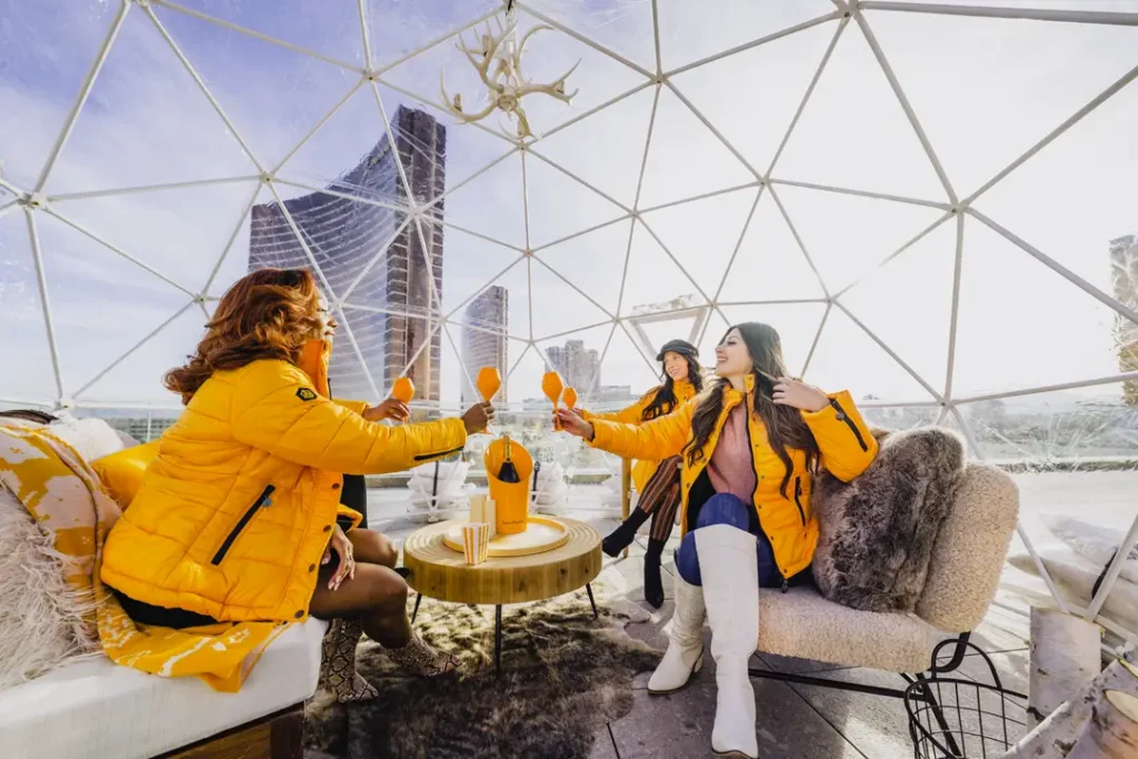 Three women in yellow jackets inside an igloo dome at Resorts World Las Vegas, raising orange glasses for a toast with a view of the city skyline.