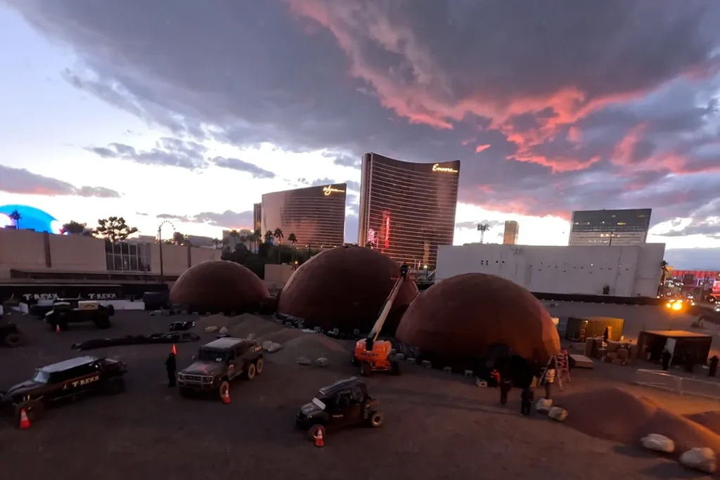 Aerial view of the Travis Scott Cactus Jack Domes event in Las Vegas at sunset. The scene showcases several large geodesic domes in an outdoor setup, surrounded by off-road vehicles and construction equipment. In the background, the Wynn and Encore hotels are illuminated against a dramatic sky filled with vibrant pink and purple clouds, creating a striking contrast with the urban landscape.