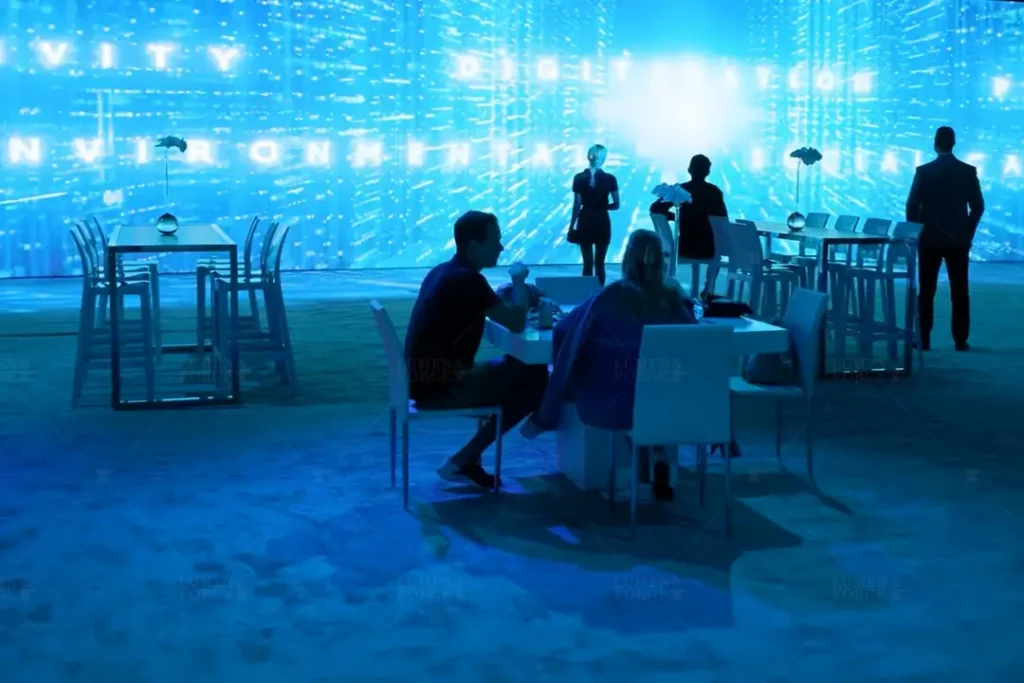 A futuristic Toyota tradeshow setup featuring immersive projection mapping. The space is illuminated in a vibrant blue hue with digital text and graphics displayed on a large screen. Attendees are seen seated at modern tables, engaging in discussions, while others stand and explore the environment. The high-tech ambiance enhances the interactive experience, showcasing Toyota's innovation in digital and environmental advancements.