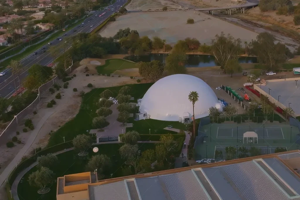 Aerial view of a large geodesic dome at an outdoor event space, surrounded by greenery, pathways, and parked vehicles.